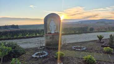 A serene countryside scene at sunset features a white statue in a brick shrine, surrounded by small plants and circular stone-bordered garden beds. Peaceful atmosphere.