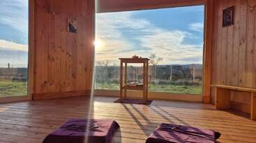 A serene wooden meditation room with large window, two purple cushions, small central table, bench, and beautiful sunlight streaming in over peaceful outdoor scenery.