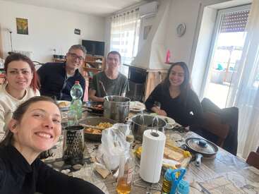 Five people are sitting around a table, smiling and enjoying a meal together in a bright, cozy dining room with food and kitchen items spread out.