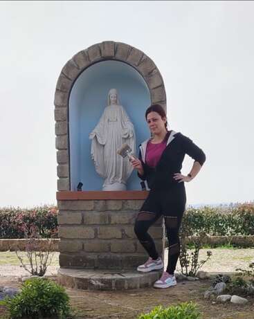 A woman in sporty attire stands beside a small outdoor shrine with a statue of Mary, holding a paintbrush, appearing to be renovating or painting it.