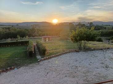 Uma bela cena campestre ao pôr do sol, com um gramado verde, cercas vivas bem aparadas, um pequeno galpão, árvores, área de cascalho e colinas ao fundo.