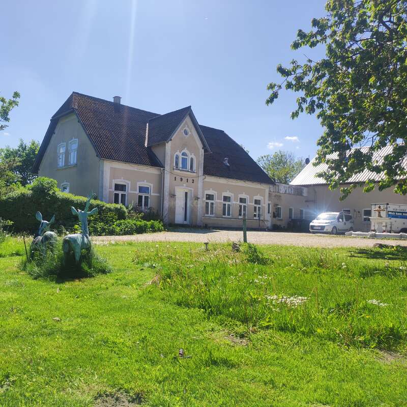 Ein charmantes Landhaus mit rotem Dach steht inmitten üppiger Vegetation, vor einem strahlend grünen Rasen, unter blauem Himmel, mit geparkten Autos und verspielten Tierskulpturen davor.