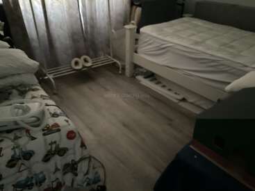 This image shows a dimly lit bedroom with two beds, a window with curtains, some bedding, a small rack, and wooden flooring, appearing cozy.
