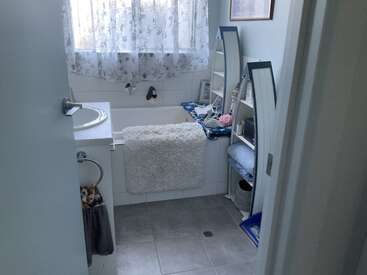 This bathroom features a bathtub with a fluffy white mat, a sink, gray tiled floor, decorative shelves with mirrors, and floral curtains, creating a cozy atmosphere.