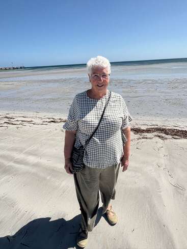 An elderly woman with white hair stands smiling on a sandy beach. She wears glasses, a checkered shirt, loose pants, and carries a patterned crossbody bag.