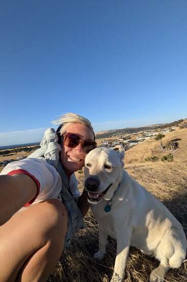 Une femme portant des lunettes de soleil serre dans ses bras son chien blanc tout sourire tandis qu'elle prend un selfie sur une colline herbeuse et aride surplombant une ville, sous un ciel bleu limpide.