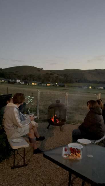 À la tombée de la nuit, un groupe de personnes est assis autour d'un foyer extérieur et admire la vue sur la campagne. Une table garnie de fruits et de collations se trouve à proximité. L'ambiance est chaleureuse.