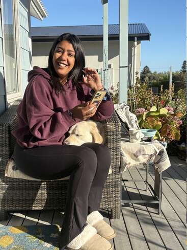 Une femme vêtue d'un sweat à capuche bordeaux sourit, assise sur une chaise de terrasse, son téléphone à la main, tandis qu'un chien repose sa tête sur ses genoux. Une matinée détendue.