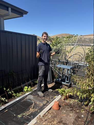 Un jeune homme vêtu de gants noirs et de vêtements sombres se tient dans un jardin ensoleillé, souriant et levant le pouce, entouré de plantes, de pots et d'outils de jardinage.