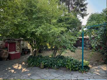 The image depicts a serene outdoor setting, featuring a stone pathway, a red water fountain, and lush greenery, with a rustic stone building in the background.