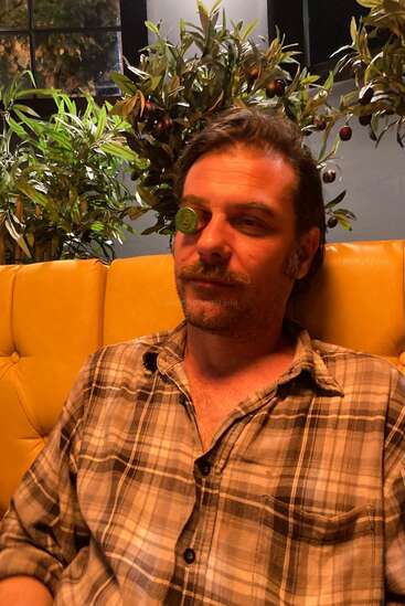 A man in a plaid shirt sits on a yellow couch, with a green coin or token covering his right eye, surrounded by leafy plants indoors.