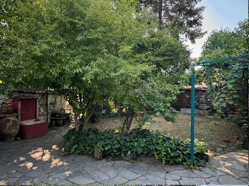 The image depicts a serene outdoor setting, featuring a stone pathway, a red water fountain, and lush greenery, with a rustic stone building in the background.