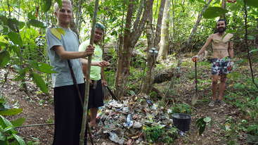 Drei Personen stehen in einer Reihe, umgeben von einem Müllhaufen, um den herum eine große Menge Abfall verstreut ist. Die Szene findet vor einer natürlichen Kulisse statt.