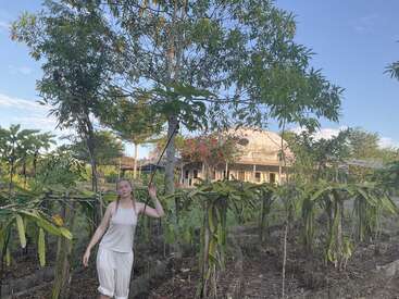 Eine Frau in weißer Kleidung steht inmitten von üppigem Grün und Drachenfruchtpflanzen in der Nähe eines rustikalen Hauses mit einem kuppelförmigen Dach unter einem blauen Himmel.