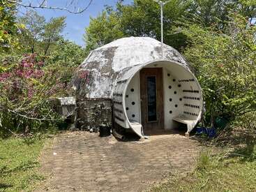 Ein kuppelförmiges, umweltfreundliches Haus mit einem runden Eingang und Bänken, umgeben von üppigem Grün und blühenden Pflanzen, mit eingelassenen Glasflaschen als Dekoration.
