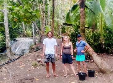 Drei Personen stehen draußen in einem tropischen Wald, umgeben von üppigem Grün und Palmen. Zwei schwarze Eimer stehen auf dem Boden, was auf eine Aktivität im Freien hindeutet.