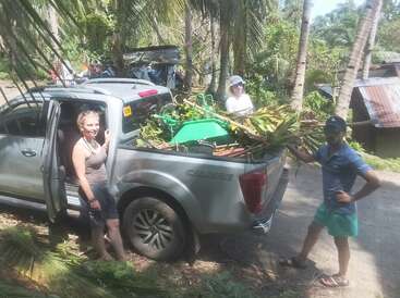 Drei Personen stehen um einen silbernen Pickup, der mit grünen Blättern, Pflanzen und Werkzeugen beladen ist. Sie scheinen sich im Freien in einer tropischen ländlichen Gegend aufzuhalten.