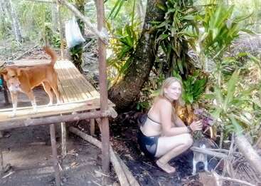 Eine Frau im schwarzen Badeanzug hockt in der Nähe von tropischen Pflanzen und hält Blumen. Ein brauner Hund steht auf einer Bambusplattform, während ein anderer kleiner Hund in der Nähe ist.