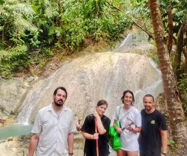 Vier Menschen posieren glücklich vor einem malerischen Wasserfall, umgeben von üppiger grüner tropischer Vegetation, und genießen ein Outdoor-Abenteuer inmitten der Natur.