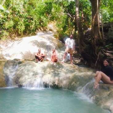 Quatro pessoas desfrutam de um dia ensolarado em uma piscina natural, sentadas e em pé sobre as rochas com água em cascata, cercadas por uma floresta tropical verdejante. Ambiente descontraído.