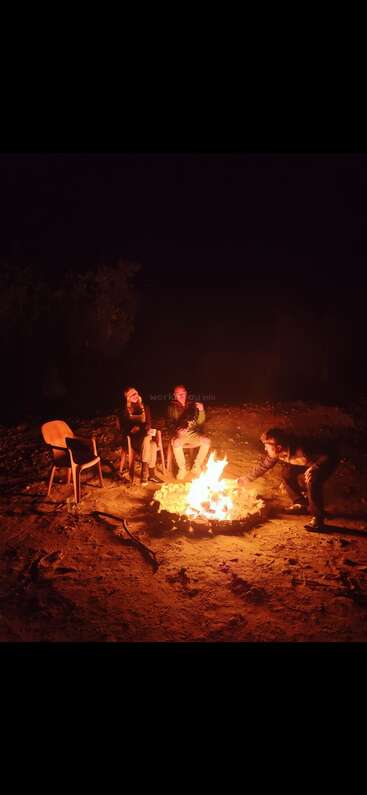 Three people sit around a campfire at night, enjoying each other’s company. One person tends the fire, while the others relax on chairs, chatting.