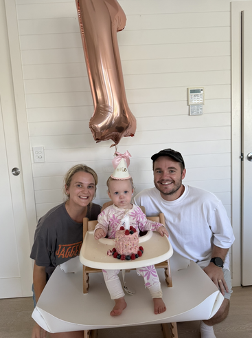 Ein Baby, das seinen ersten Geburtstag feiert, sitzt mit einem rosa Kuchen in einem Hochstuhl, trägt einen Partyhut und ist von zwei lächelnden Erwachsenen und einem großen Luftballon umgeben.