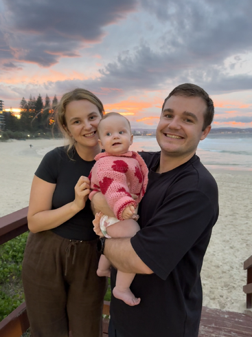 A happy family of three poses on a beach at sunset. The parents smile while holding their baby in a pink sweater with red hearts.
