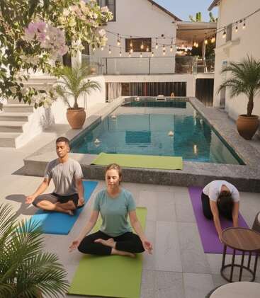 Three people practice yoga on colorful mats beside a serene pool, surrounded by potted plants, string lights, peaceful ambiance, and white buildings under clear blue sky.