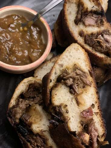 Sliced bread stuffed with tender, juicy meat sits on a dark plate, accompanied by a small bowl of thick, rich brown sauce with a spoon.