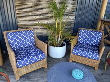 Two wicker armchairs with blue patterned cushions sit around a round table. A potted palm plant stands between the chairs; bamboo and metal wall behind.