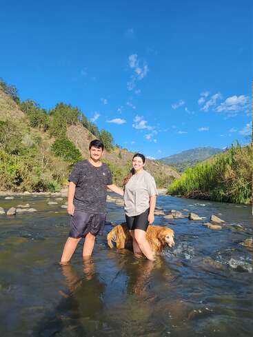 Ein Mann und eine Frau stehen lächelnd in einem seichten, klaren Fluss mit einem Golden Retriever. Üppig grüne Hügel und blauer Himmel bilden eine schöne, sonnige Kulisse.