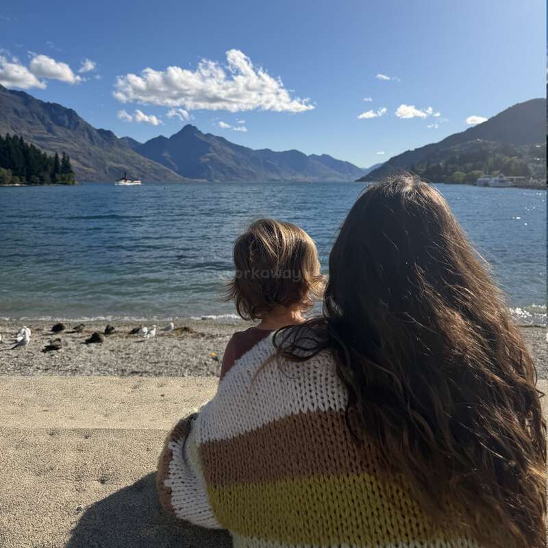 Eine Frau und ein Kind sitzen am Sandstrand und schauen auf einen ruhigen See, der von Bergen umgeben ist und unter einem klaren blauen Himmel liegt. Sie genießen einfach die Schönheit der Natur.