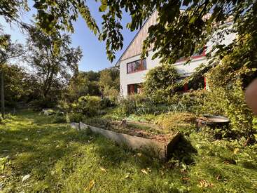 Um jardim exuberante circunda uma casa branca com janelas com guarnições vermelhas, muita vegetação, árvores e luz do sol. Dá pra ver canteiros elevados e um carrinho de mão.