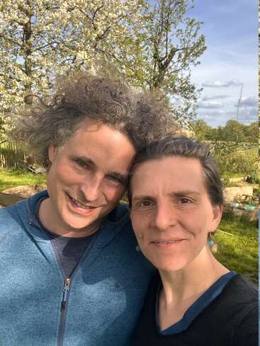 Um casal sorridente está bem juntinho ao ar livre, sob um céu azul, com árvores floridas e uma vegetação exuberante ao fundo, aproveitando um dia ensolarado na natureza.