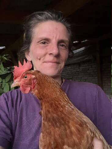 Uma mulher vestindo uma blusa roxa sorri calorosamente para a câmera enquanto segura uma galinha marrom. O fundo mostra um cenário rústico com construções de tijolo e madeira.