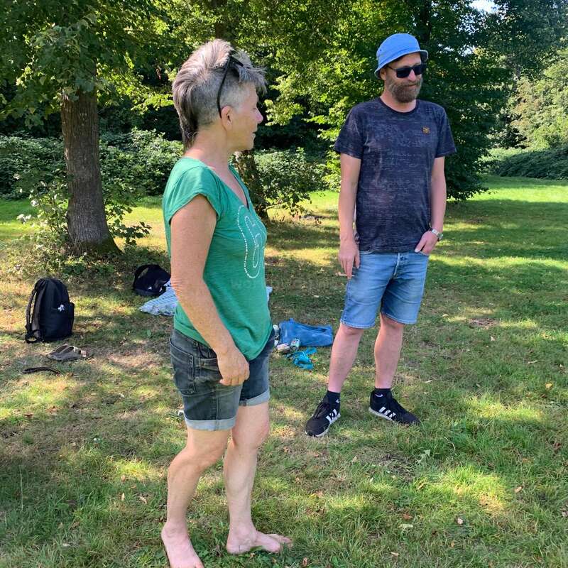 Una mujer y un hombre conversan sobre la hierba de un parque soleado. Llevan ropa informal de verano, rodeados de árboles y mochilas.