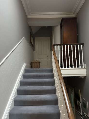 Una escalera alfombrada con barandilla de madera conduce a un pasillo superior. Arriba se ven una cesta de mimbre, cuadros enmarcados y un armario alto.