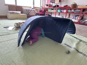 A young child crawls through a tunnel made of a folded mat on a padded floor in a playroom filled with toys and shelves of supplies.
