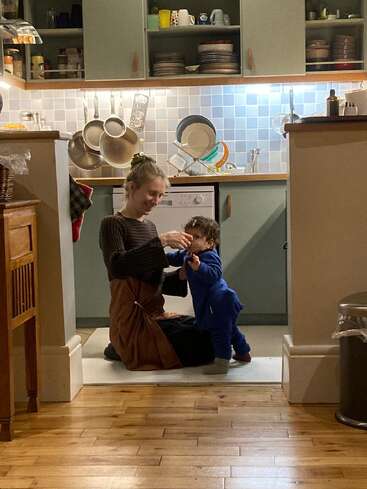 A woman kneels on the kitchen floor, smiling and helping a young child walk. The cozy kitchen background features hanging pans and dishes on a drying rack.