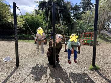 Une femme est agenouillée entre deux enfants assis sur des balançoires dans une aire de jeux. Le soleil illumine le parc ; on aperçoit des jeux et des arbres en arrière-plan. Les visages ne sont pas visibles.
