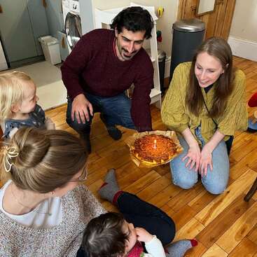 A group of adults and children gather around a birthday cake with a candle, smiling and celebrating together in a cozy home setting with wooden floors.