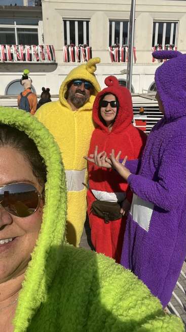 Cuatro adultos disfrazados de Teletubbies posan juntos al aire libre, con trajes de peluche de colores, gafas de sol y una sonrisa en un día soleado, junto a un edificio de aspecto festivo.