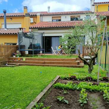 A backyard garden with vegetable beds, green lawn, a small tree, wooden deck, outdoor seating, trampoline, and a modern house with tiled roof in background.