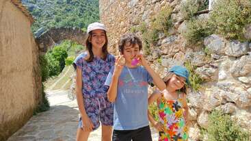 Três crianças estão em um caminho de pedra ensolarado ao lado de um muro antigo, vestindo roupas coloridas de verão e chapéus, sorrindo e aproveitando um dia ensolarado e alegre ao ar livre.