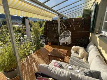 Un acogedor patio exterior con suelo de madera, una silla colgante, plantas en macetas, un cómodo sofá, una mesa de centro y un perro mullido tomando el sol.