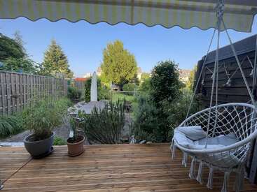 Una acogedora terraza de madera con macetas, un columpio colgante y un toldo amarillo da a un tranquilo y verde jardín con árboles, valla y muebles de exterior.