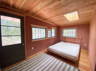 Dieses gemütliche Schlafzimmer besticht durch holzvertäfelte Decken, ein Oberlicht, Wände in Erdtönen, blau umrandete Fenster, ein minimalistisches Bett, einen gestreiften Teppich und viel Tageslicht.