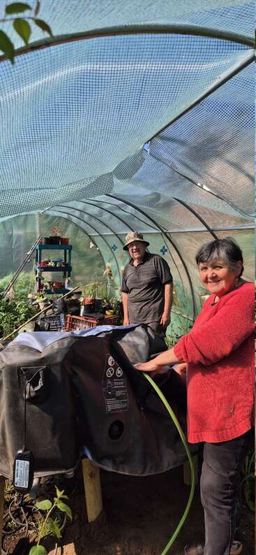 Eine Frau in einem roten Pullover gießt Pflanzen in einem grünen Gewächshaus. Ein lächelnder Mann steht in der Nähe zwischen Gartengeräten, Töpfen und üppiger Vegetation.