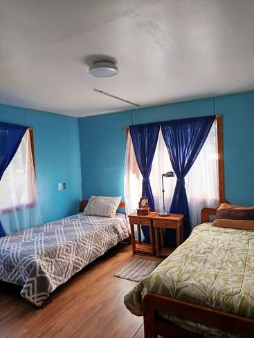 Dieses gemütliche Schlafzimmer hat zwei Einzelbetten mit gemusterten Steppdecken, blaue Wände, durchsichtige Vorhänge mit dunklen Paneelen, einen hölzernen Nachttisch, eine Lampe und einen Holzfußboden. Natürliches Licht erfüllt den Raum.