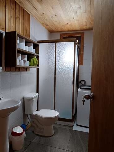 Dieses Badezimmer hat eine Holzdecke, eine Dusche mit Milchglas, eine weiße Toilette und ein Waschbecken, Regale mit Toilettenpapier, eine Waschmaschine und einen kleinen Mülleimer.